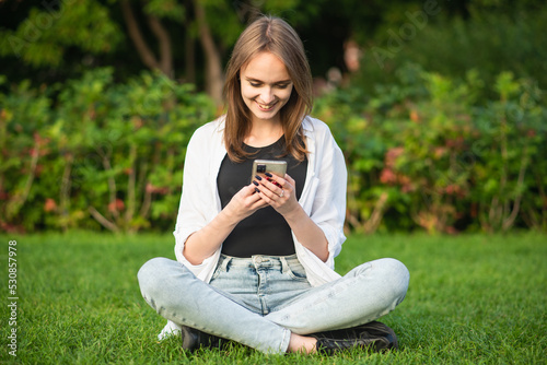 A young and attractive Caucasian girl in casual clothes is sitting on the grass in the park and using a mobile phone.