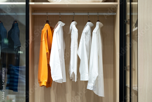 Shirts hang in a walking closet with glass door. One of those is in orange standout from others that are in white.