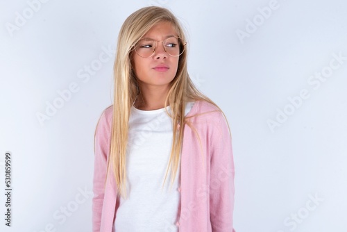 caucasian blonde little girl wearing pink jacket and glasses over white background   looks pensively aside  plans actions after university  imagines what to do Thinks over about new project.