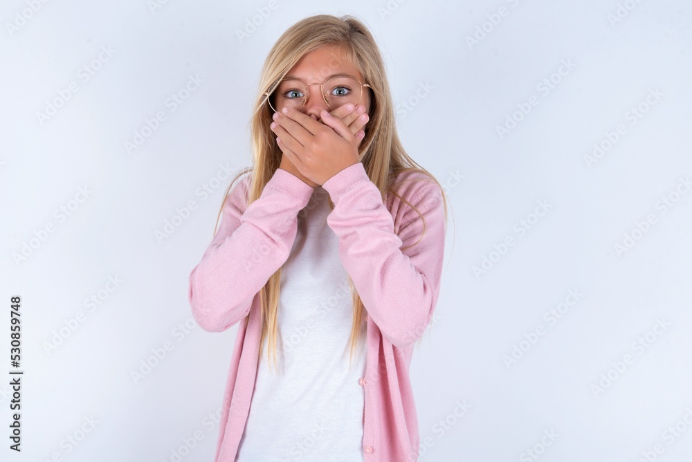 Upset caucasian blonde little girl wearing pink jacket and glasses over white background , covering her mouth with both palms to prevent screaming sound, after seeing or hearing something bad.