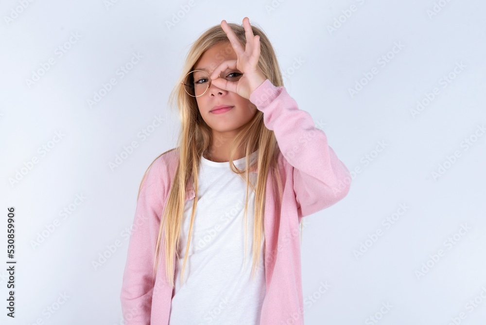 caucasian blonde little girl wearing pink jacket and glasses over white background with happy face smiling doing ok sign with hand on eye looking through finger.