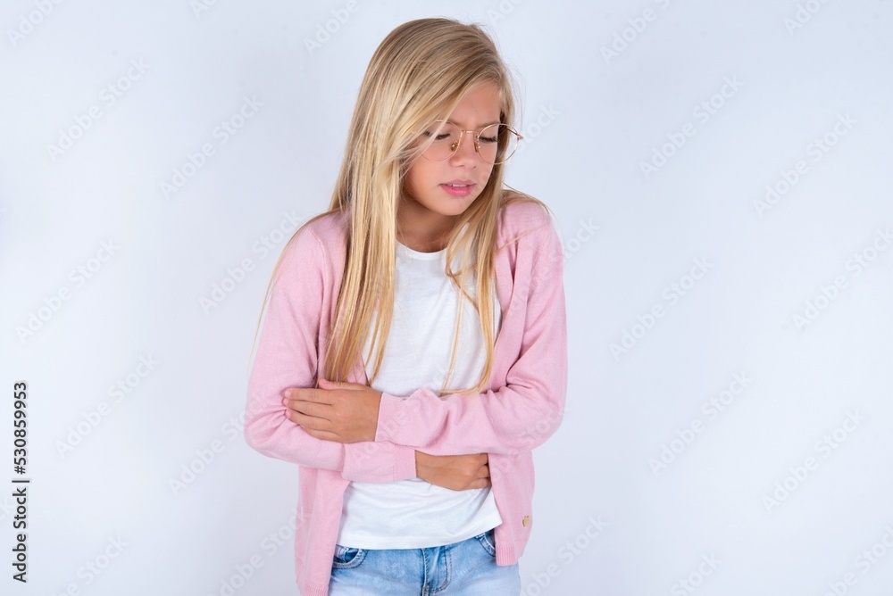 caucasian blonde little girl wearing pink jacket and glasses over white background suffering from strong stomachache. caucasian blonde little girl wearing pink jacket and glasses over white background suffering from strong stomachache.