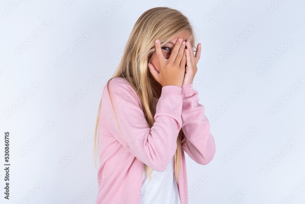 caucasian blonde little girl wearing pink jacket and glasses over white background covering face with hands and peering out with one eye between fingers. Scared from something or someone. caucasian blonde little girl wearing pink jacket and glasses over white background covering face with hands and peering out with one eye between fingers. Scared from something or someone.