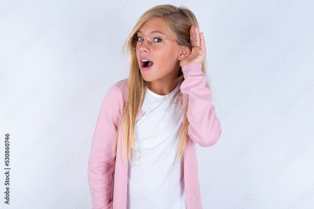 Oh my God! Funny astonished caucasian blonde little girl wearing pink jacket and glasses over white background opening mouth widely looking aside, with hand near ear trying to listen to gossips. Oh my God! Funny astonished caucasian blonde little girl wearing pink jacket and glasses over white background opening mouth widely looking aside, with hand near ear trying to listen to gossips.