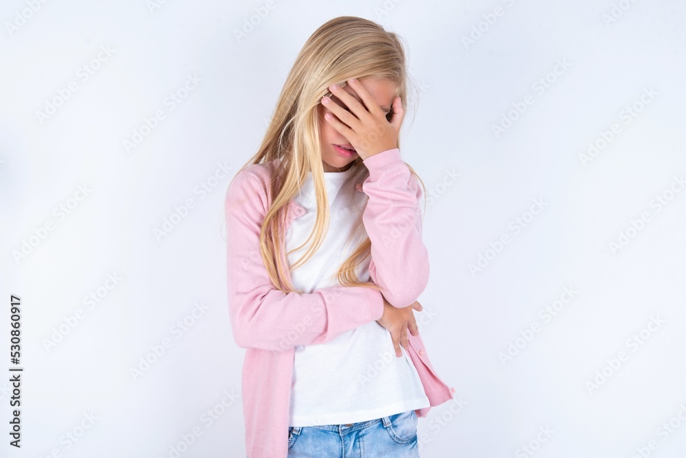 caucasian blonde little girl wearing pink jacket and glasses over white background making facepalm gesture while smiling amazed with stupid situation. caucasian blonde little girl wearing pink jacket and glasses over white background making facepalm gesture while smiling amazed with stupid situation.