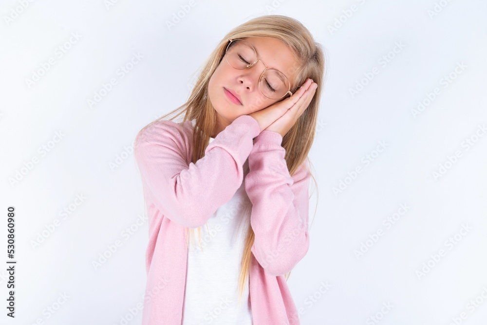 Relax and sleep time. Tired caucasian blonde little girl wearing pink jacket and glasses over white background with closed eyes leaning on palms making sleeping gesture. Relax and sleep time. Tired caucasian blonde little girl wearing pink jacket and glasses over white background with closed eyes leaning on palms making sleeping gesture.