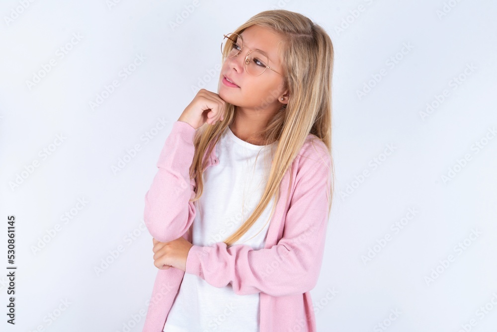 Face expressions and emotions. Thoughtful caucasian blonde little girl wearing pink jacket and glasses over white background holding hand under his head, having doubtful look.