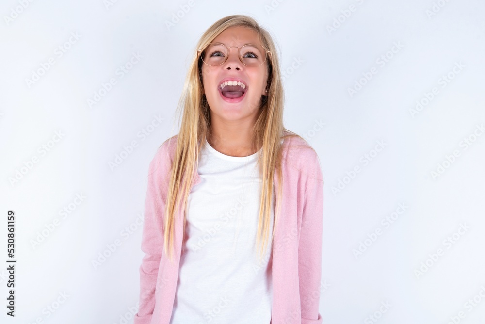 Surprised caucasian blonde little girl wearing pink jacket and glasses over white background , shrugs shoulders, looking sideways, being happy and excited. Sudden reactions concept. Surprised caucasian blonde little girl wearing pink jacket and glasses over white background , shrugs shoulders, looking sideways, being happy and excited. Sudden reactions concept.