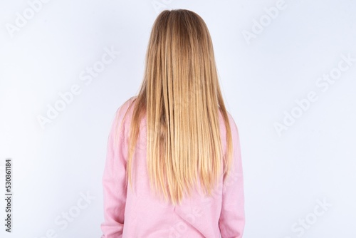 caucasian blonde little girl wearing pink jacket and glasses over white background standing backwards looking away with arms on body.