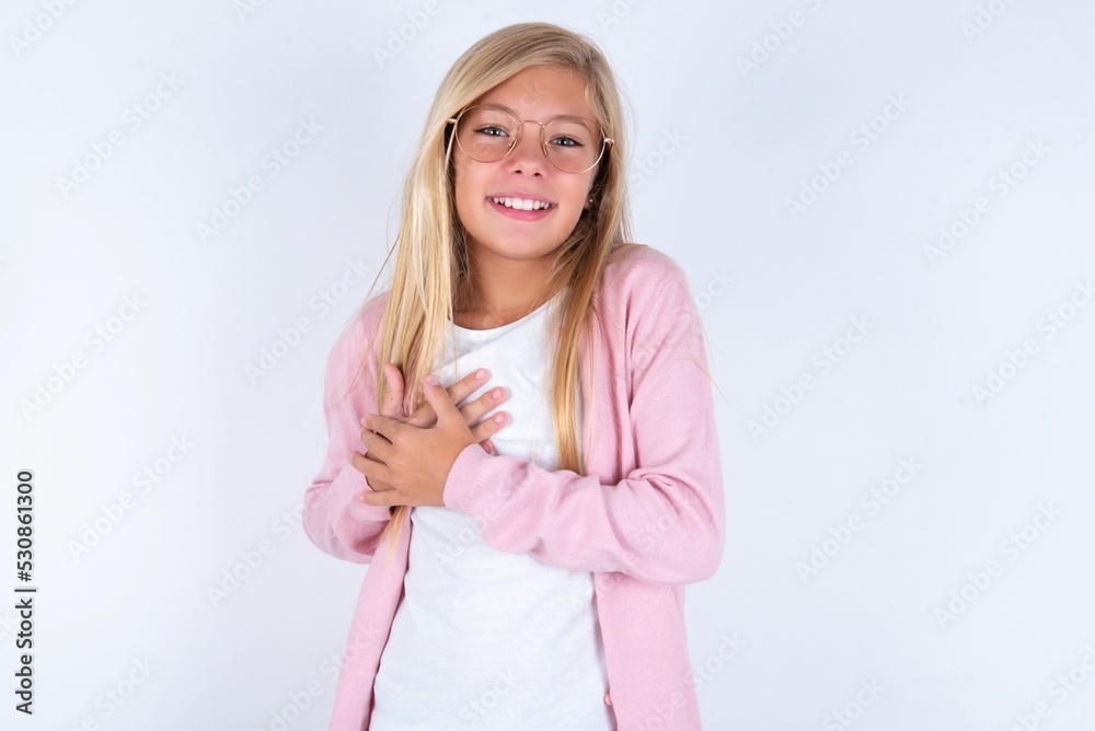 Honest caucasian blonde little girl wearing pink jacket and glasses over white background keeps hands on chest, touched by compliment or makes promise, looks at camera with great pleasure. Honest caucasian blonde little girl wearing pink jacket and glasses over white background keeps hands on chest, touched by compliment or makes promise, looks at camera with great pleasure.
