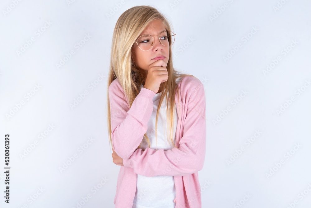 caucasian blonde little girl wearing pink jacket and glasses over white background with hand under chin and looking sideways with doubtful and skeptical expression, suspect and doubt. caucasian blonde little girl wearing pink jacket and glasses over white background with hand under chin and looking sideways with doubtful and skeptical expression, suspect and doubt.