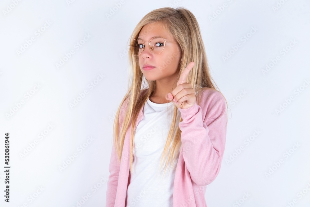 No sign gesture. Closeup portrait unhappy caucasian blonde little girl wearing pink jacket and glasses over white wall raising fore finger up saying no. Negative emotions facial expressions, feelings. No sign gesture. Closeup portrait unhappy caucasian blonde little girl wearing pink jacket and glasses over white wall raising fore finger up saying no. Negative emotions facial expressions, feelings.