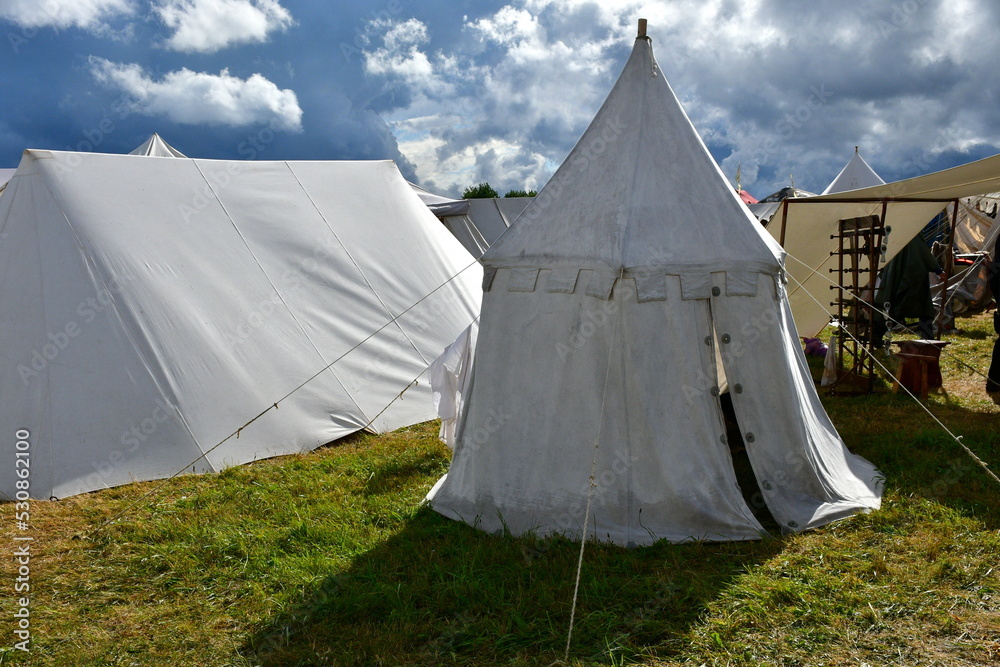 A close up on a tent made out white cloth with a wooden frame and ropes supporting it seen during a big medieval folklore festival or fair right before a massive thunderstorm and rainfall