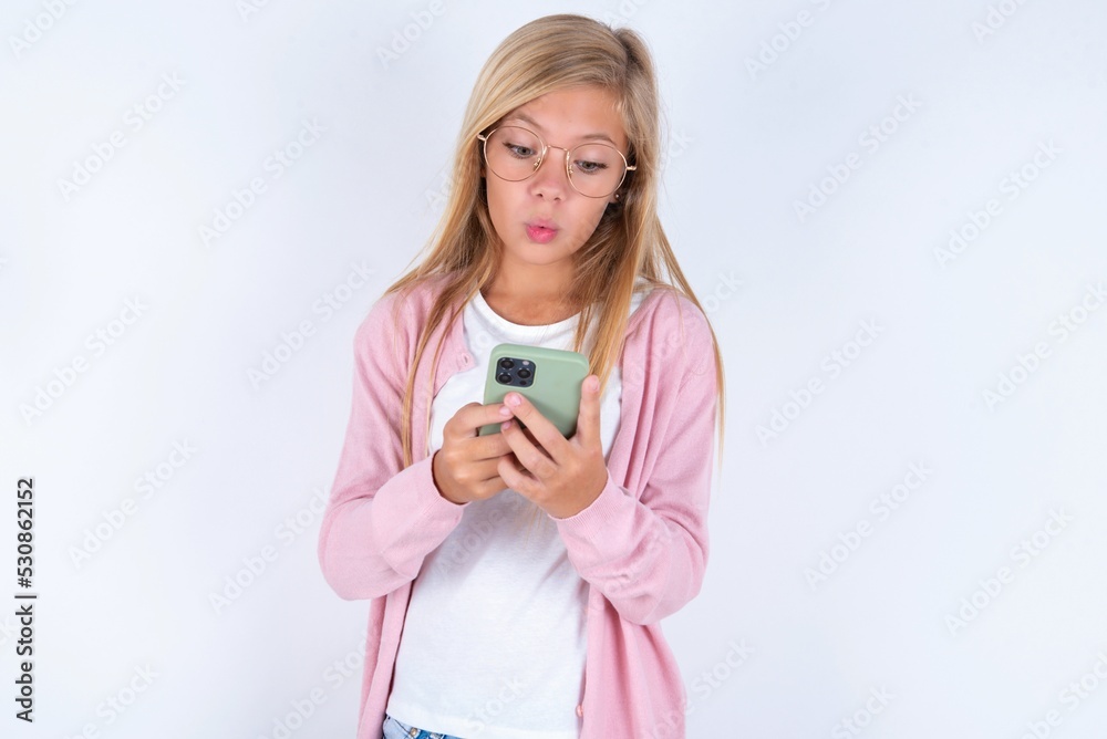 caucasian blonde little girl wearing pink jacket and glasses over white background looks with bugged eyes, holds modern smart phone, receives unexpected message from friend, reads reminder. caucasian blonde little girl wearing pink jacket and glasses over white background looks with bugged eyes, holds modern smart phone, receives unexpected message from friend, reads reminder.