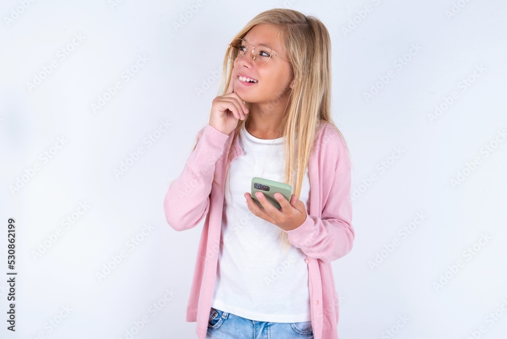 Image of a thinking dreaming caucasian blonde little girl wearing pink jacket and glasses over white background using mobile phone and holding hand on face. Taking decisions and social media concept. Image of a thinking dreaming caucasian blonde little girl wearing pink jacket and glasses over white background using mobile phone and holding hand on face. Taking decisions and social media concept.