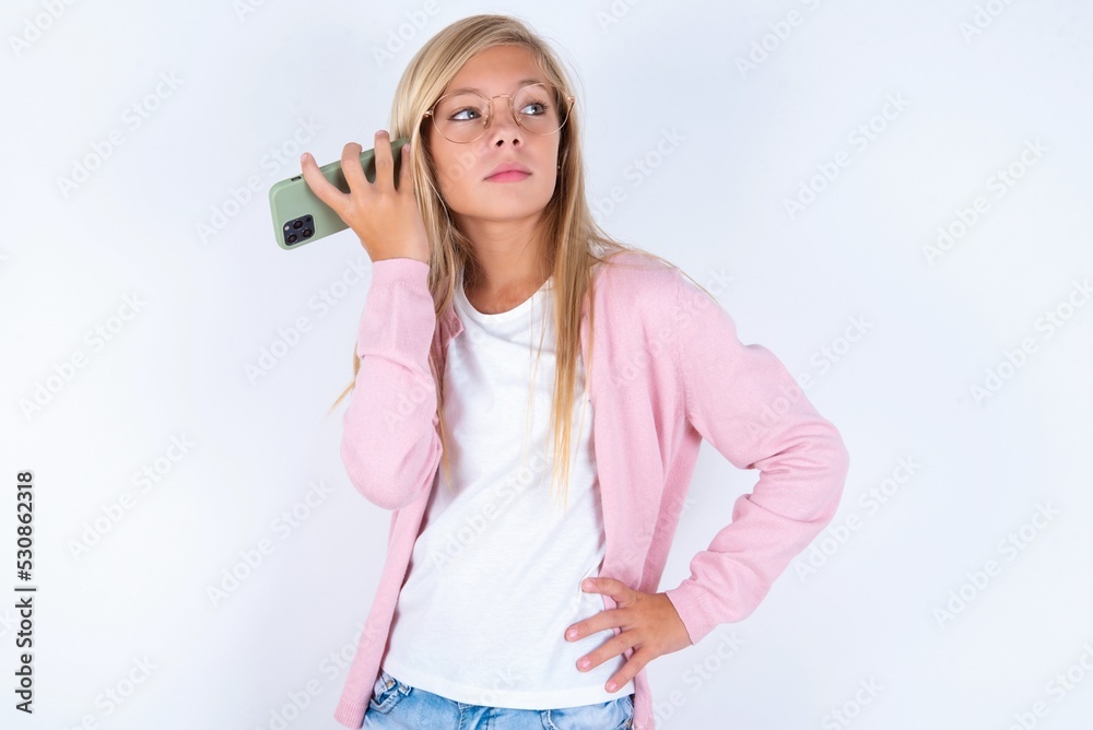 Smiling caucasian blonde little girl wearing pink jacket and glasses over white background listening a voice message from her smartphone. Communication and technology concept. Smiling caucasian blonde little girl wearing pink jacket and glasses over white background listening a voice message from her smartphone. Communication and technology concept.