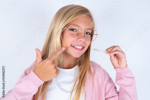 caucasian blonde little girl wearing pink jacket and glasses over white background holding an invisible aligner and pointing to her perfect straight teeth. Dental healthcare and confidence concept.