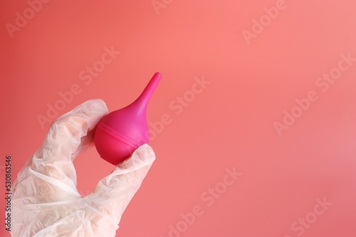 A pink enema is held by a hand in a white glove. Medical device. Pink background