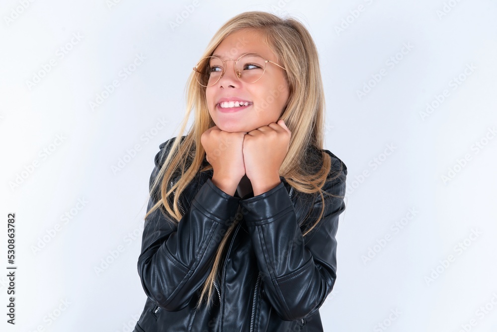 beautiful caucasian blonde little girl wearing biker jacket and glasses over white background holds hands under chin, glad to hear heartwarming words from stranger beautiful caucasian blonde little girl wearing biker jacket and glasses over white background holds hands under chin, glad to hear heartwarming words from stranger
