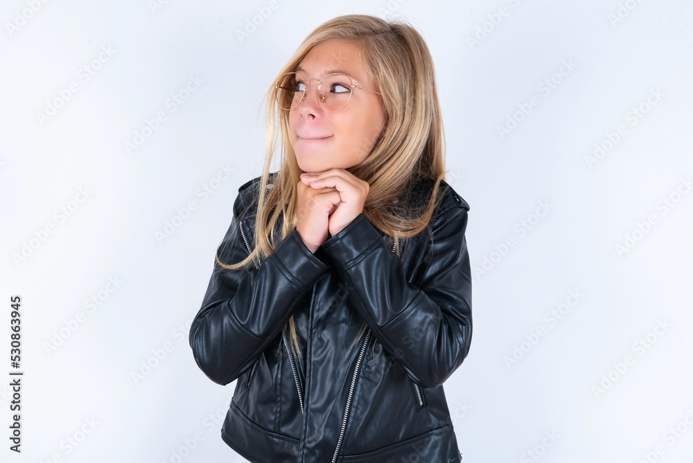 Curious beautiful caucasian blonde little girl wearing biker jacket and glasses over white background keeps hands under chin bites lips and looks with interest aside. Curious beautiful caucasian blonde little girl wearing biker jacket and glasses over white background keeps hands under chin bites lips and looks with interest aside.