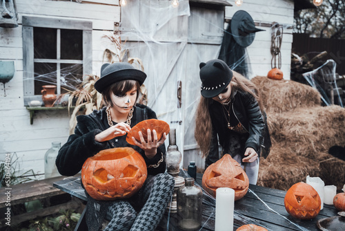 Beautiful scary little girls celebrating halloween. Terrifying black, white half-face makeup,witch costume, stylish image.Fun at children's party in barn on street. Hat, jacket,pumpkin jack-o-lantern