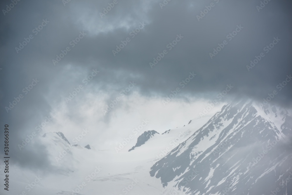 Atmospheric mountain landscape with fuzzy silhouettes of rocks on glacier in low clouds during rain. Dramatic view to large glacier tongue in snow mountains blurred in rain haze in gray low clouds.