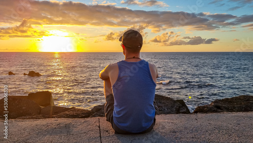 Tourist man with hat and tank top sitting at port of Puerto de la Cruz during sunset  Tenerife  Canary Islands  Spain  Europe. Vacation at the sea. Reflection of sun beams in the calm sea surface