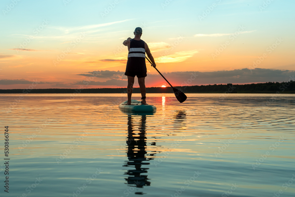 A man in shorts on a sapboard with an oar against the backdrop of a sunset sky swims in the lake in the evening.