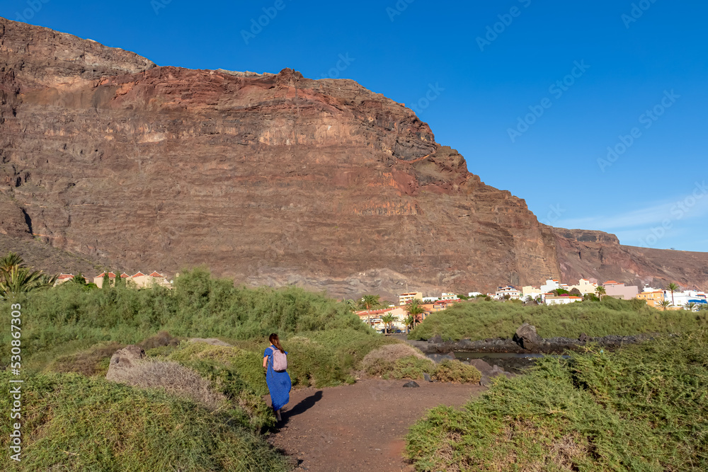 Woman in blue dress walking along shoreline with panoramic view on massive sharp cliffs and mountains in village Valle Gran Rey, La Gomera, Canary Islands, Spain, Europe. Beach Playa Charco del Conde