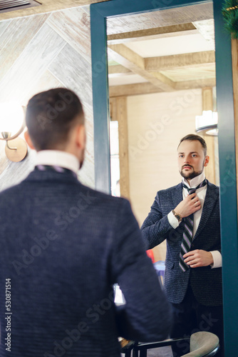 A young man with a small beard adjusts his tie while looking in a large mirror. Elegantly dressed young man.