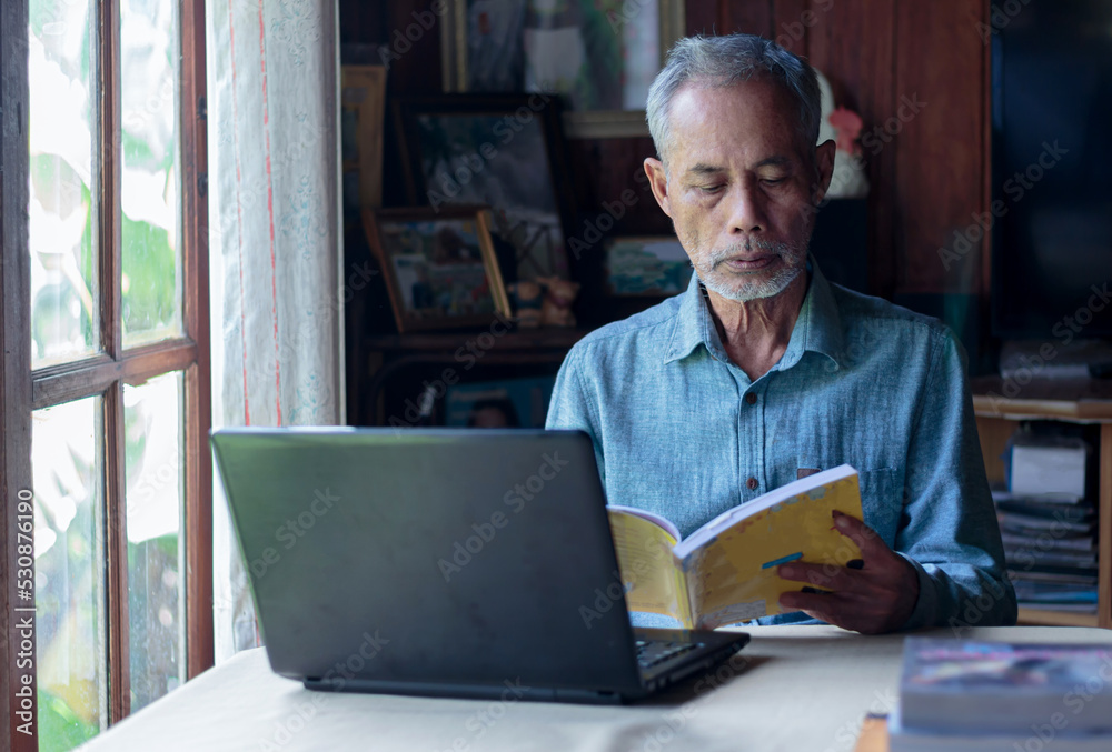 Asian senior man reading a book using a notebook senior lifestyle concept Asian senior man reading a book using a notebook senior lifestyle concept