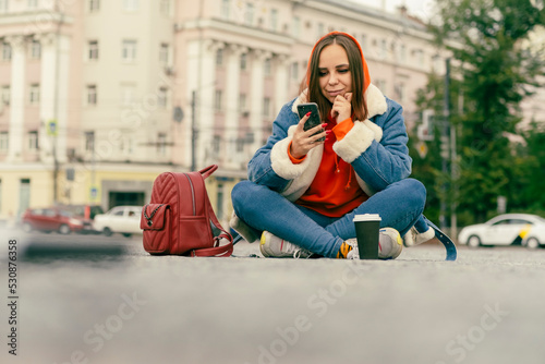 Beautiful young woman sitting outside on street together with cell phone Outdoors city background. Lifestyle