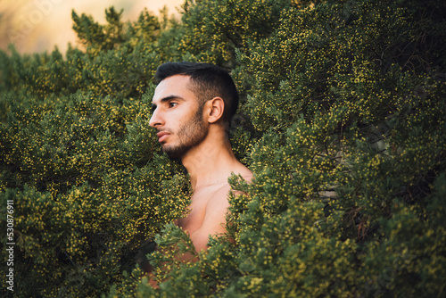 Shirtless ethnic guy standing amidst green bushes and looking away