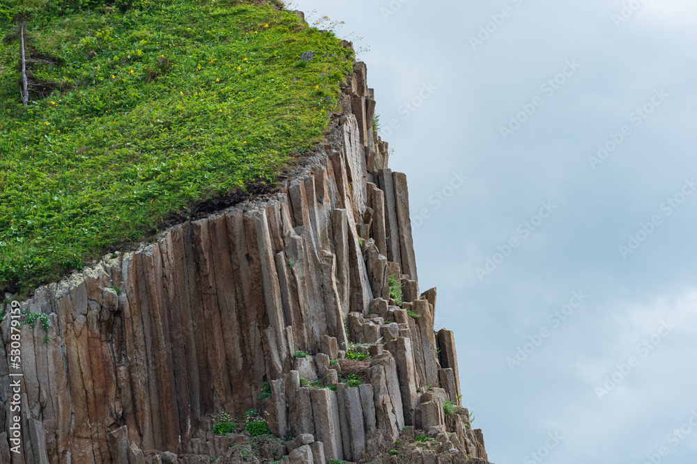 Fototapeta premium fragment of a cliff formed by columnar basalt with lush grass on top against the sky