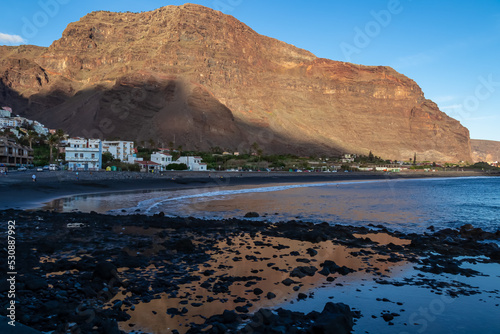 Scenic view on beach Playa Valle Gran Rey during sunset seen from Promenade La Calera in Valle Gran Rey on La Gomera, Canary Islands, Spain, Europe. Mountain reflections in dark volcanic sand beach