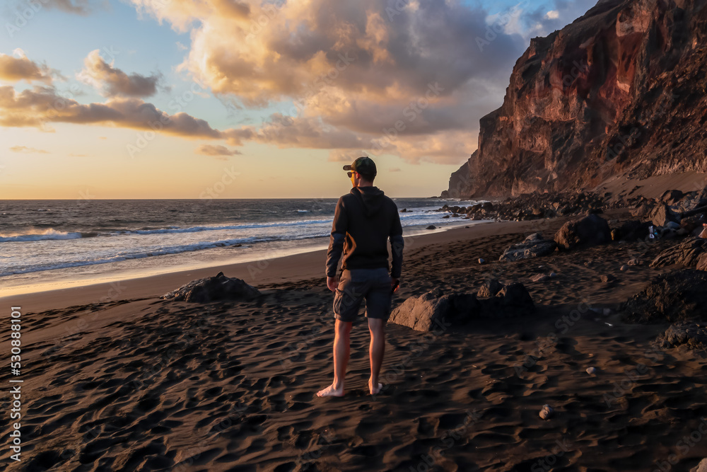 Woman in dress walking barefoot on volcanic sand beach Playa del Ingles during sunset in Valle Gran Rey on La Gomera, Canary Islands, Spain, Europe. Calm atmosphere seaside. Massive cliffs La Mercia