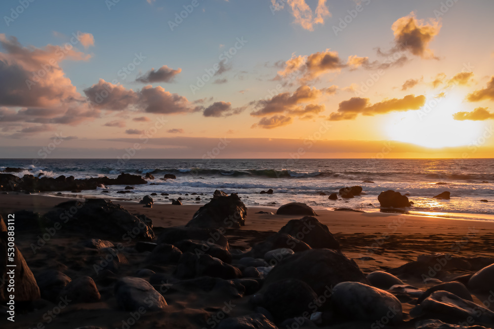 Scenic view during sunset on the volcanic sand beach Playa del Ingles in Valle Gran Rey, La Gomera, Canary Islands, Spain, Europe. Sun beam reflection on water surface. Stones in the foreground