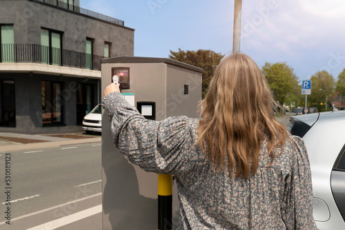 Eine Frau steht an einer E-Auto Ladestation. Sie bezahlt kontaktlos. Im Hintergrund ist eine Straße mit Verkehr. photo