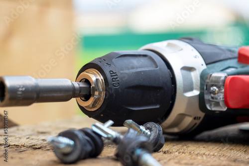 Close-up of a cordless screwdriver lying on a wooden surface with a nozzle for roofing screws. Self-tapping screws painted in gray are lying nearby. selective focus