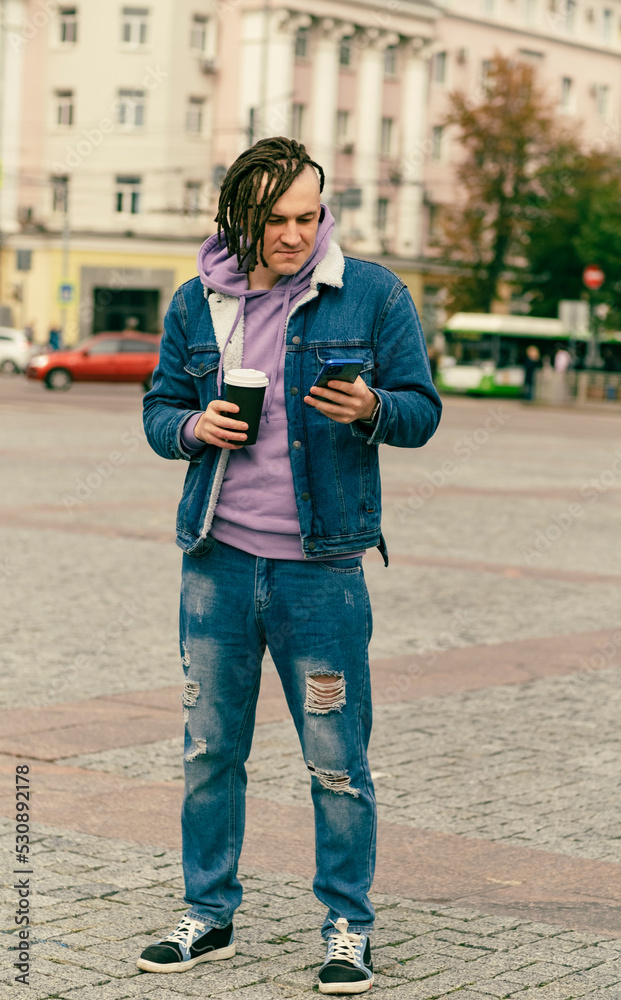 Hipster man with dreadlocks drinking hot coffee on street together with cell phone Outdoors city background. Young male in warm clothes drinking hot beverage