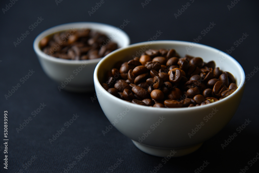 Delicious coffee beans in a ceramic white bowl on a black background. Coffee beans close-up in the dishes. Delicious coffee beans in a ceramic white bowl on a black background. Coffee beans close-up in the dishes.