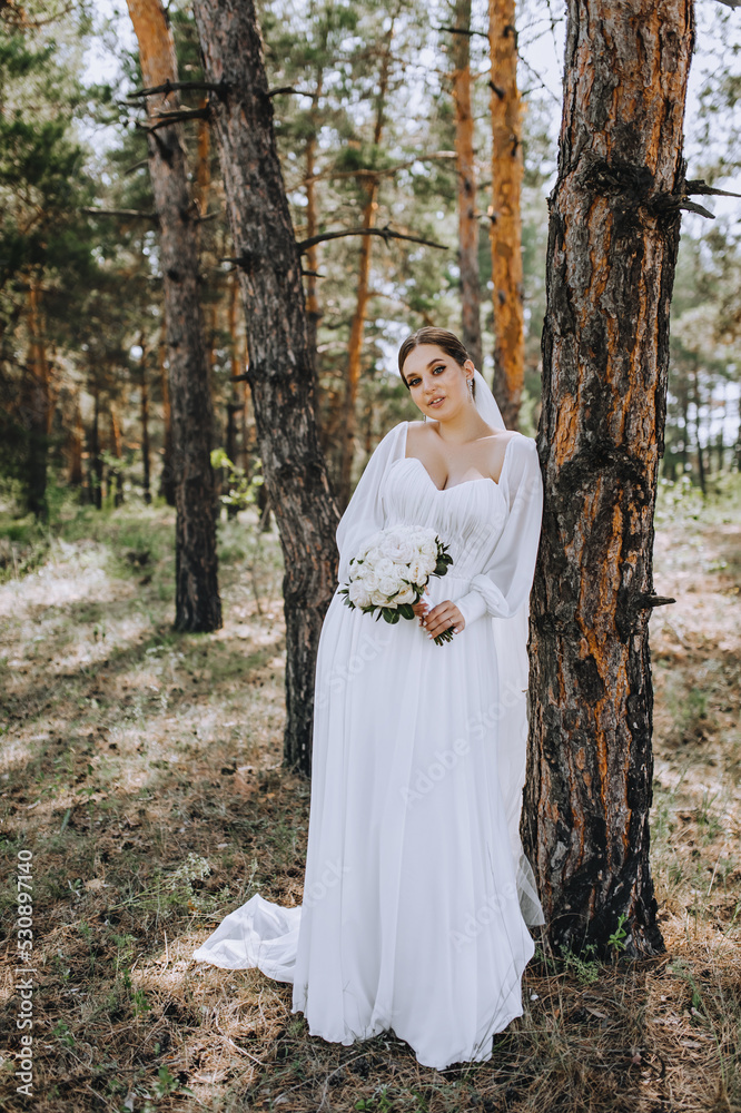 A beautiful brunette bride in a white long dress with a bouquet of roses stands in a coniferous forest in nature near a tree. Wedding portrait, photography. A beautiful brunette bride in a white long dress with a bouquet of roses stands in a coniferous forest in nature near a tree. Wedding portrait, photography.