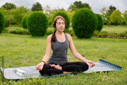 Mindful fit sporty healthy calm young woman sitting in lotus pose on mat doing yoga exercise breathing fresh air meditating in park feel balance and no stress free concept, copy space.