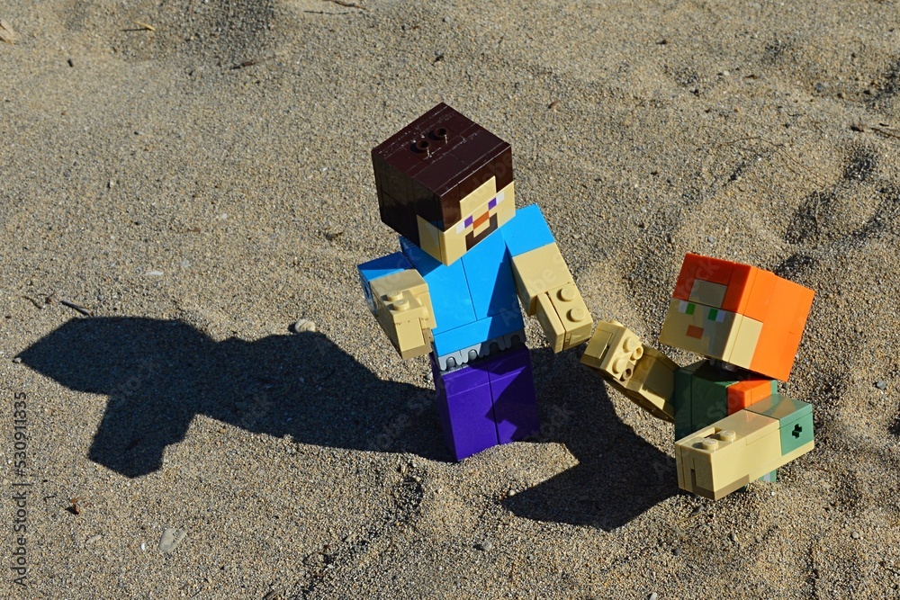 Fototapeta premium LEGO Minecraft large action figure of Steve in blue shirt pulling out of desert shifty sand pit his female friend Alex. Summer afternoon sunshine. 