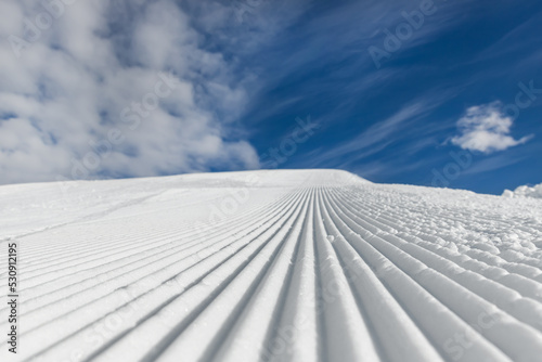 Close-up straight line rows of freshly prepared groomed ski slope piste with bright shining sun and clear blue sky background. Snowcapped mountain downhill landscape at europe winter skiing resort