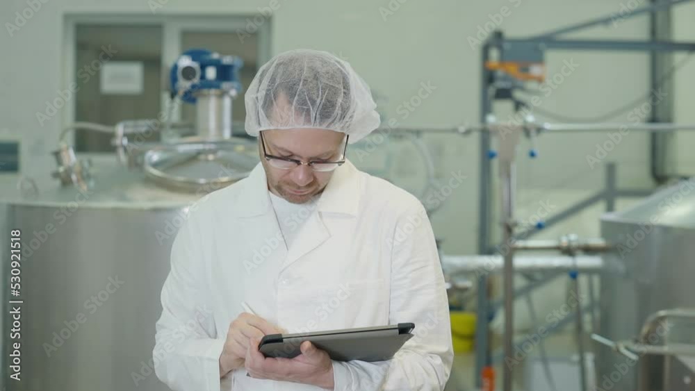 Factory worker holding a tablet in his hands. Factory worker wearing white clothes using a portable tablet. Factory worker presses multiple buttons on the tablet touchscreen with a stylus. Factory worker holding a tablet in his hands. Factory worker wearing white clothes using a portable tablet. Factory worker presses multiple buttons on the tablet touchscreen with a stylus.