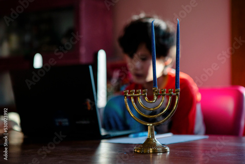 A boy sits at table on a computer, a menorah in foreground photo
