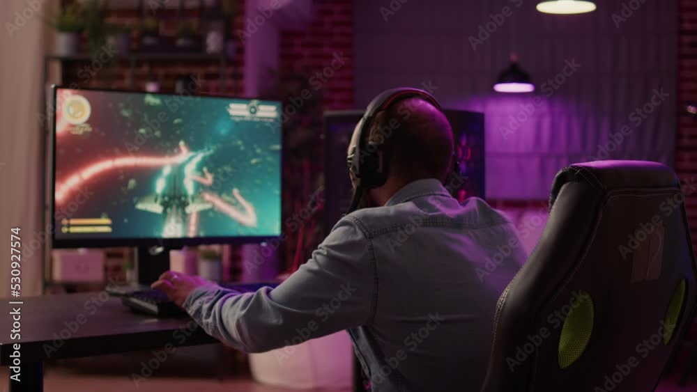 Over shoulder view of caucasian man using pc setup playing multiplayer online action game talking to team on headset. Gamer streaming fast paced space shooter while explaining gameplay to subscribers.