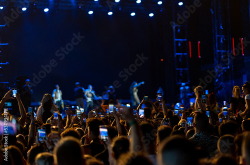 Closeup rear view of large crowd of people enjoying an open air concert on a summer night. There are many raised hands holding smart phones and taping the show instead of actually watching it.