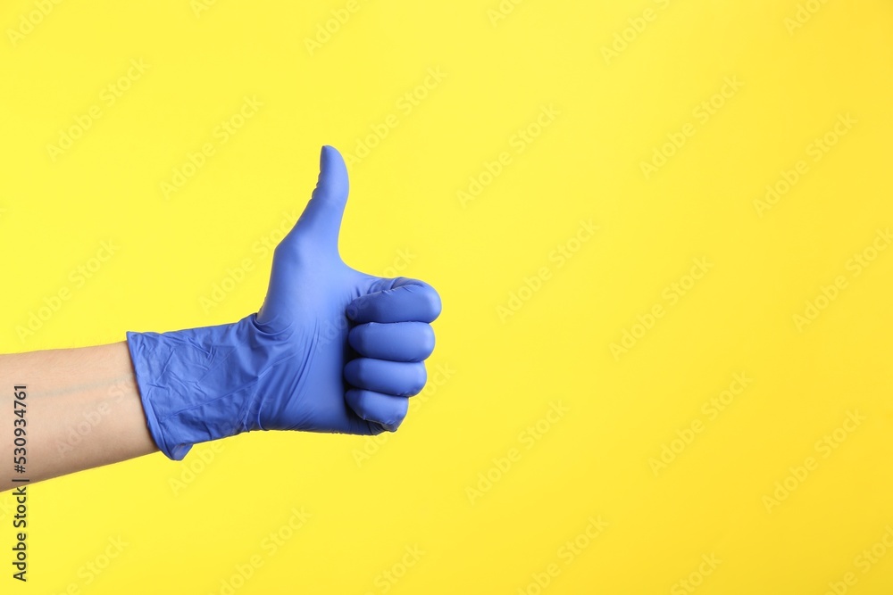 Person in medical gloves showing thumb up on yellow background, closeup of hand. Space for text